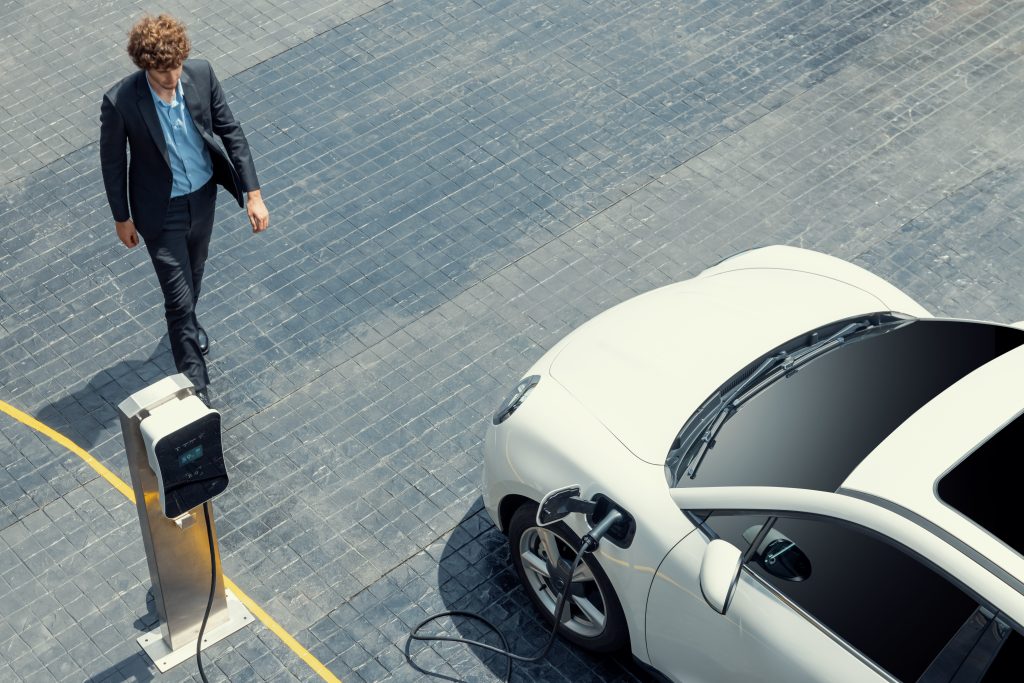 Aerial view of a businessman in black formal suit with his electric vehicle recharging battery at public car park charging station.